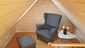 Una habitación pequeña y acogedora con un sillón tapizado de color gris, un otoman a juego y una pequeña mesa redonda con un jarrón de flores blancas.