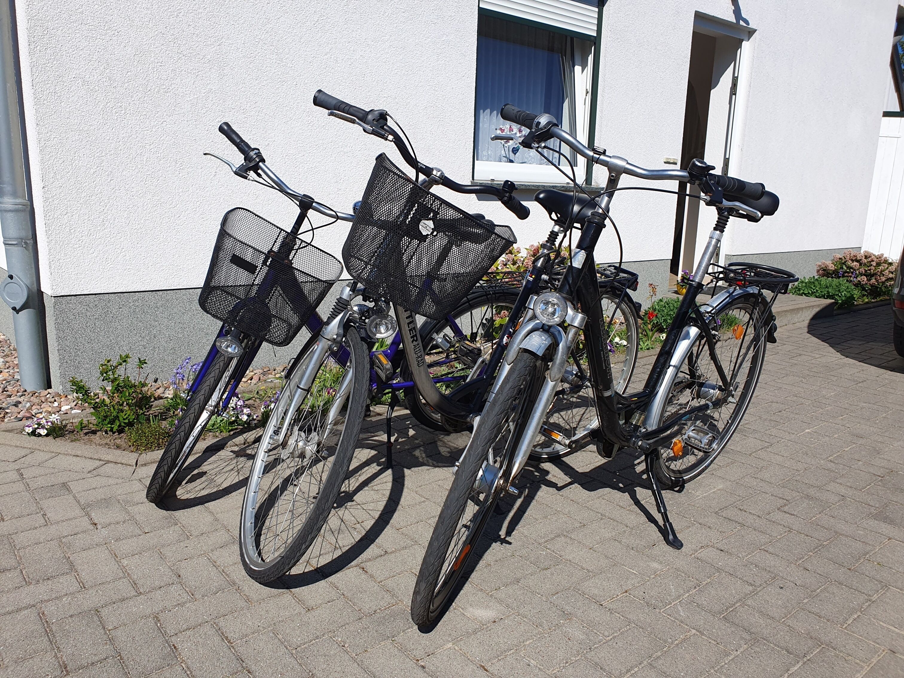 Deux vélos sont garés sur une zone pavée devant un bâtiment à la façade blanche, avec une fenêtre ornée d’un rideau bleu.