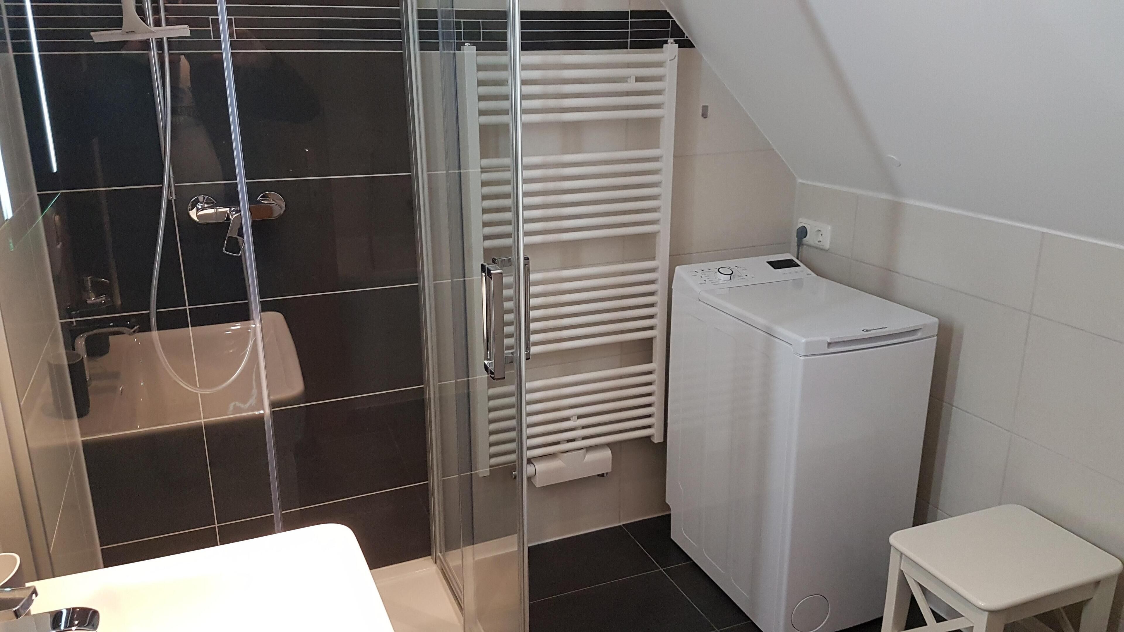 A modern bathroom with a glass shower enclosure, a white towel rack, a built-in washing machine, and a small white stool.