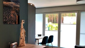 Une cuisine moderne avec un plan de travail en bois, un réfrigérateur blanc et un tableau représentant une scène de plage. On y trouve un coin repas avec deux chaises et une table, ainsi qu’une grande fenêtre donnant sur un paysage verdoyant et un bâtiment.