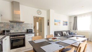 A modern kitchen with a built-in oven, a dining area with a table and chairs, and a living area with a sofa and cushions. - Beachfront 3-room apartment for 5 people with WiFi and wallbox (Graal-Müritz)