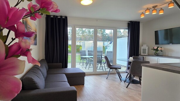 A modern living room with a grey sofa, a dining area with a table and chairs, and a kitchen area with a TV and a vase of flowers. - 2-zi.-fewo für 3 Pers. im EG und Gartenanteil (Graal-Müritz)