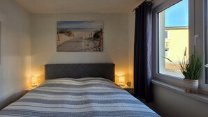 A bedroom with a bed, two bedside lamps, a window with a view of a building, and a painting of a lighthouse on the wall. - 2-zi.-fewo für 3 Pers. im EG und Gartenanteil (Graal-Müritz)