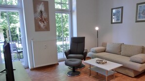 A modern living room with a sofa, armchair, and coffee table. There is a radiator, a painting on the wall, and a window with a view of trees. - 2-room apartment for 3 people close to the beach (Graal-Müritz)