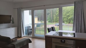 Ein modernes Wohnzimmer mit Flachbildfernseher, Sofa, Esstisch und Küchenzeile inklusive Backofen. Große Fenster bieten einen Blick ins Freie.