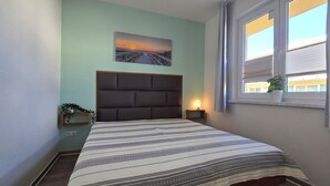 Un dormitorio con cama, mesita de noche, ventana con cortinas, una planta colgada en la pared y un cuadro con un atardecer sobre un puente.
