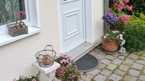 Una casa amb una porta blanca i finestres, un petit porxo amb plantes en testos i un jardí ple de flors diverses.