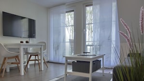 Ein modernes Wohnzimmer mit Flachbildfernseher, weißem Schreibtisch, zwei Stühlen, einem kleinen Tisch mit Korb und großen Fenstern mit Jalousien.
