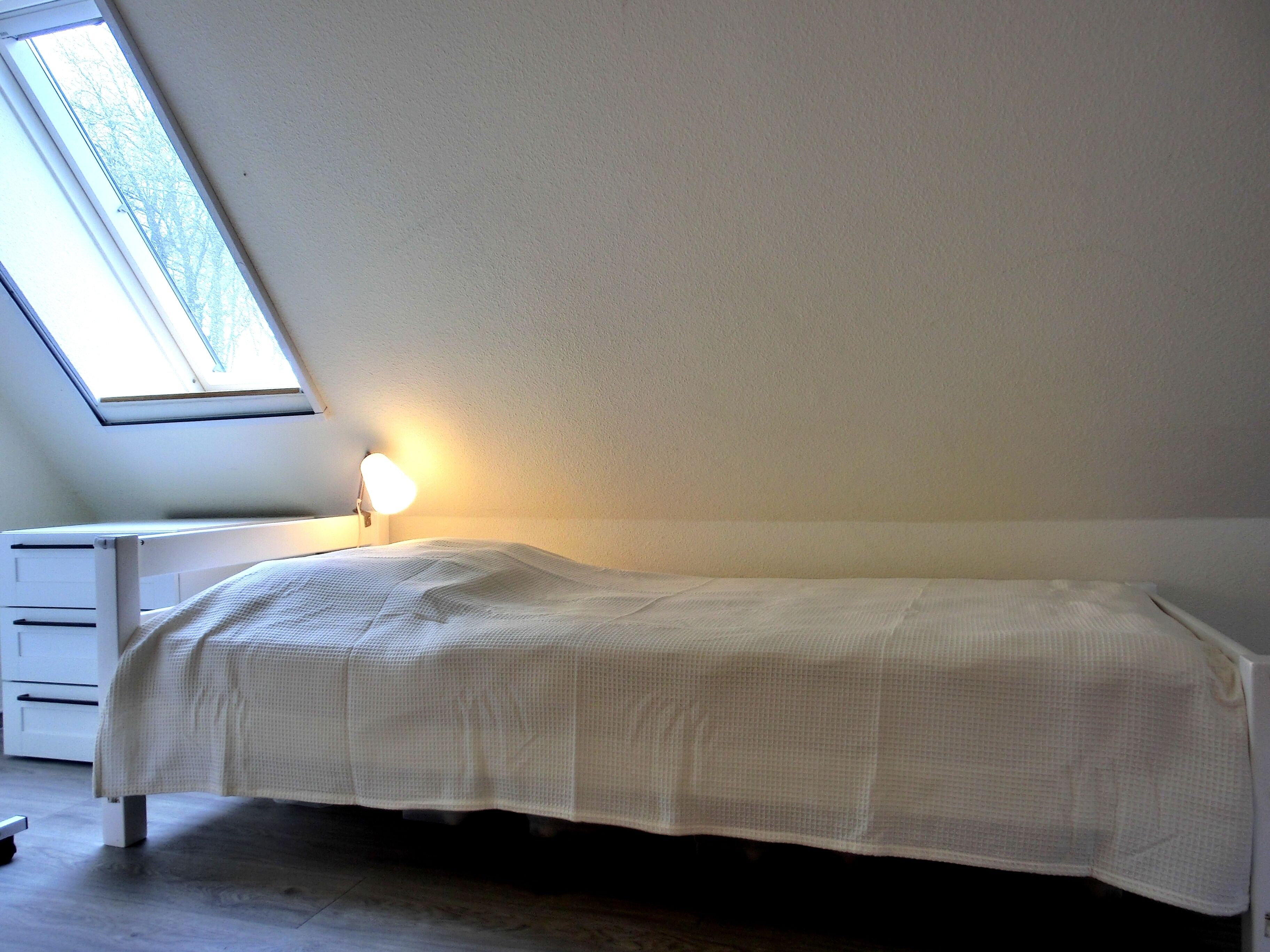 Habitación pequeña con cama individual, tragaluz y lámpara de noche.