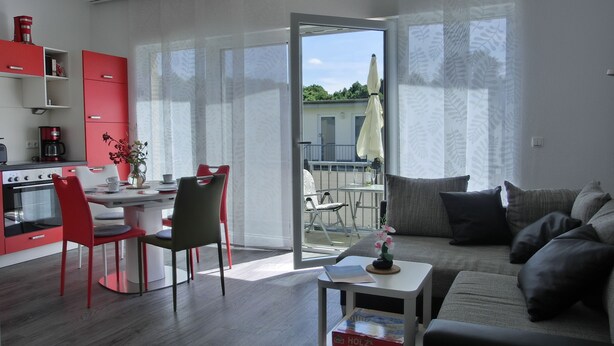 Ein modernes Wohnzimmer mit einem grauen Sofa, einem Couchtisch mit einem Buch und einem Essbereich mit roten Stühlen und einem weißen Tisch.