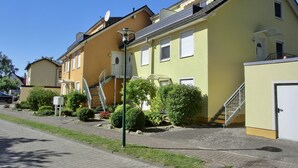 A yellow building with a staircase leading to a balcony, surrounded by greenery and a paved area. - Große, Komfortable 3 Zimmer-maisonette-ferienwohnung für 5 Personen (Graal-Müritz)