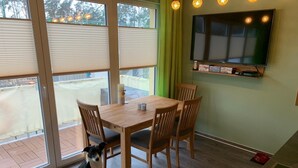 Een eetruimte met een houten tafel en stoelen, een aan de muur bevestigde televisie, en een hond die bij het raam zit.