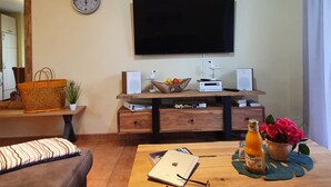 A living room with a flat-screen TV mounted on the wall, a wooden entertainment unit with speakers, a woven basket, and a bottle of orange juice on a wooden table.