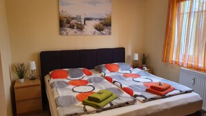 A neatly made bed with a patterned bedspread, a bedside table with a lamp and a plant, and a painting of a beach scene on the wall.
