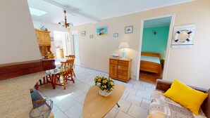 Un salon chaleureux avec une table à manger en bois, un canapé agrémenté d’un coussin jaune et un lit adossé à un mur vert. Des tableaux encadrés ornent le mur et une lampe est posée sur la table.