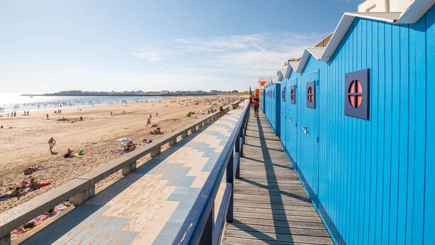 Une promenade en bois mène à une plage bordée d’une rangée de bâtiments bleus dotés de fenêtres.