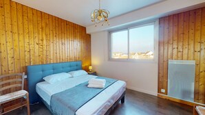 A bedroom with a wooden paneled wall, a large bed with white linens, a blue headboard, a wooden chair, a window with a view, and a heater.