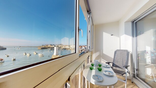 A balcony with a table set for dining, overlooking a harbor with boats and a coastal town.