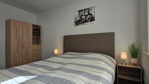 Une chambre à coucher comprenant un lit, une armoire, une table de chevet avec une lampe, un tableau accroché au mur et une fenêtre avec des stores.
