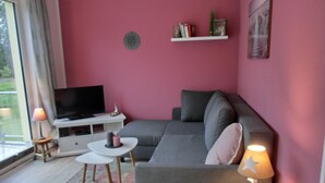 Een moderne woonkamer met een grijze bank, een klein wit tafeltje, een televisie op een witte kast, en een roze muur.