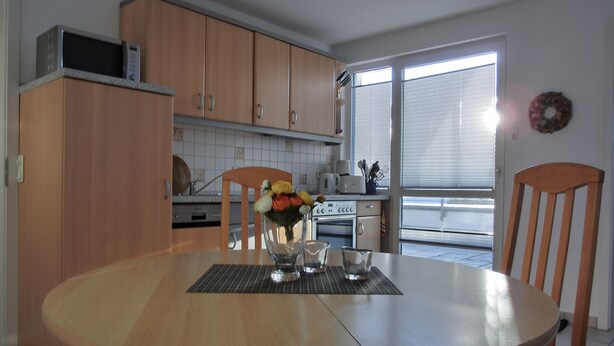 A dining area with a round table, chairs, a vase of flowers, and a kitchen in the background. - 2-zimmer-fewo für 3 Pers. im Dachgeschoss (Graal-Müritz)