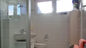 A modern bathroom with a glass shower enclosure, a toilet, and a sink. There is a window providing natural light and a towel rack with a towel. - 2-zimmer-fewo für 3 Pers. im Dachgeschoss (Graal-Müritz)