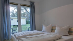 A bedroom with a bed, two pillows, a window with curtains, and a view of a building and trees outside. - Modern 2-room vacation apartment for 3 people with bicycles and WLAN (Graal-Müritz)
