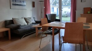 A modern living room with a sofa, a dining table with chairs, a floor lamp, and a view of a house through a glass door. - Modern 2-room vacation apartment for 3 people with bicycles and WLAN (Graal-Müritz)