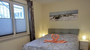 Un dormitorio con cama, ventana con persianas, cortina, mesita de noche con lámpara y una obra de arte en la pared.