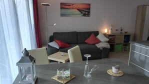 Una sala de estar moderna con un sofá gris, cojines rojos y blancos, un cuadro de un atardecer, una lámpara de pie y una pequeña mesa con una vela y cristalería.