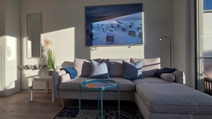 A modern living room with a grey sectional sofa, a small side table, a framed picture of a beach scene, and a large window. - 2-zimmer-fewo für 2 Pers. im OG mit 2 Fahrrädern (Graal-Müritz)