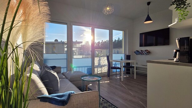 A modern living space with a sofa, dining table, and chairs, large windows with a view, and a kitchen area. - 2-zimmer-fewo für 2 Pers. im OG mit 2 Fahrrädern (Graal-Müritz)