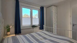 A bedroom with a bed, a window with curtains, a door with floral wallpaper, and a potted plant. - 2-zimmer-fewo für 2 Pers. im OG mit 2 Fahrrädern (Graal-Müritz)