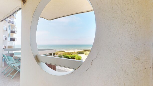 A circular window overlooking a beach with a view of the sea and a building in the background. - Le Concorde, appartement face mer (Saint-Gilles-Croix-de-Vie)