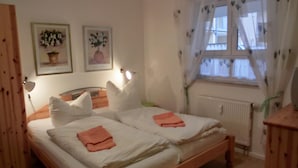 Une chambre avec deux lits, du linge de lit blanc, une tête de lit en bois, une fenêtre avec des rideaux légers et deux lampes fixées au mur.
