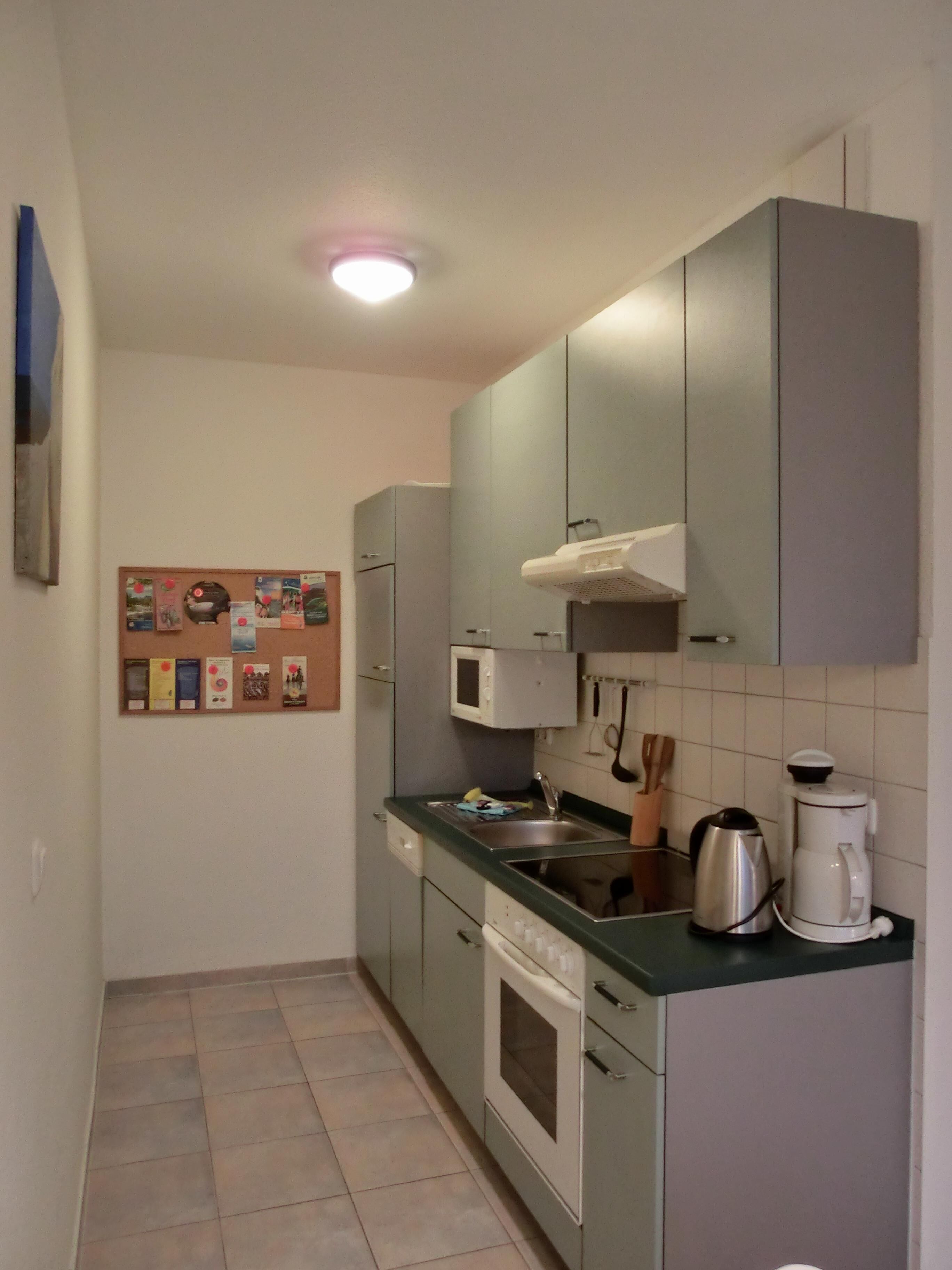 Eine kleine Küche mit Mikrowelle, Herd, Spüle und Schränken. Ein Korkbrett ist mit verschiedenen Gegenständen an der Wand befestigt.