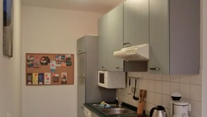 Une petite cuisine équipée d’un four à micro-ondes, d’une cuisinière, d’un évier et de placards. Un panneau d’affichage en liège est fixé au mur, sur lequel divers objets sont accrochés.