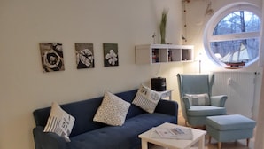 Un salon avec un canapé bleu foncé, un fauteuil bleu clair, une table basse blanche et une petite table d’appoint blanche. Trois tableaux sont accrochés au mur, ainsi qu’une étagère blanche où sont posés des livres et des objets décoratifs. Une fenêtre offre une vue sur les arbres.