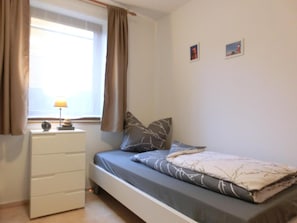 Ein Schlafzimmer mit einem Bett, einer Kommode, einem Fenster mit Vorhängen und zwei kleinen Bildern an der Wand.