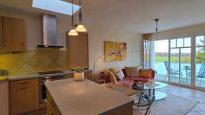 Eine moderne Küche mit Holzschränken, einem eingebauten Backofen und einer großen Kochinsel. Ein Essbereich mit einem Glastisch und Stühlen lädt zum Verweilen ein, und ein großes Fenster bietet Ausblick ins Freie.