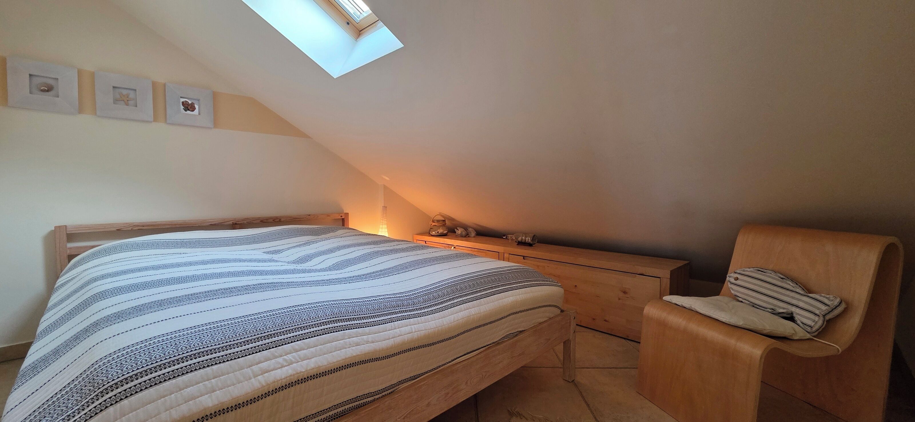 A bedroom with a sloped ceiling, a skylight, a bed with a striped bedspread, a wooden nightstand, a wooden chair, and a small dresser.