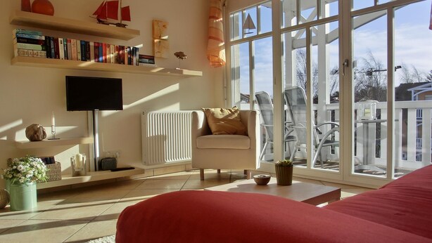 Ein Wohnzimmer mit einer Couch, einem Fernseher, einem Bücherregal und einem großen Fenster mit Blick auf Gartenmöbel.