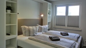 Ett sovrum med en säng, ett sängbord, en hylla med en växt, ett fönster med persienner och en taklampa.