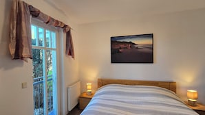 A bedroom with a bed, a nightstand with a lamp, a TV, a window with curtains, and a painting on the wall.