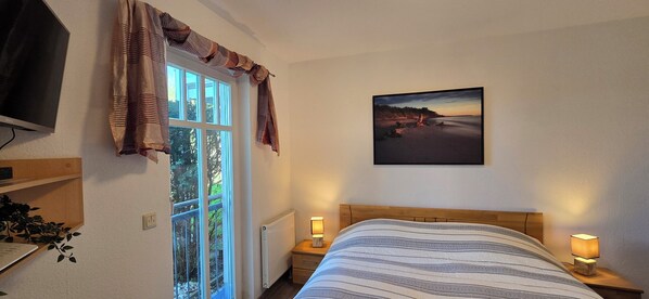A bedroom with a bed, a nightstand with a lamp, a TV, a window with curtains, and a painting on the wall.