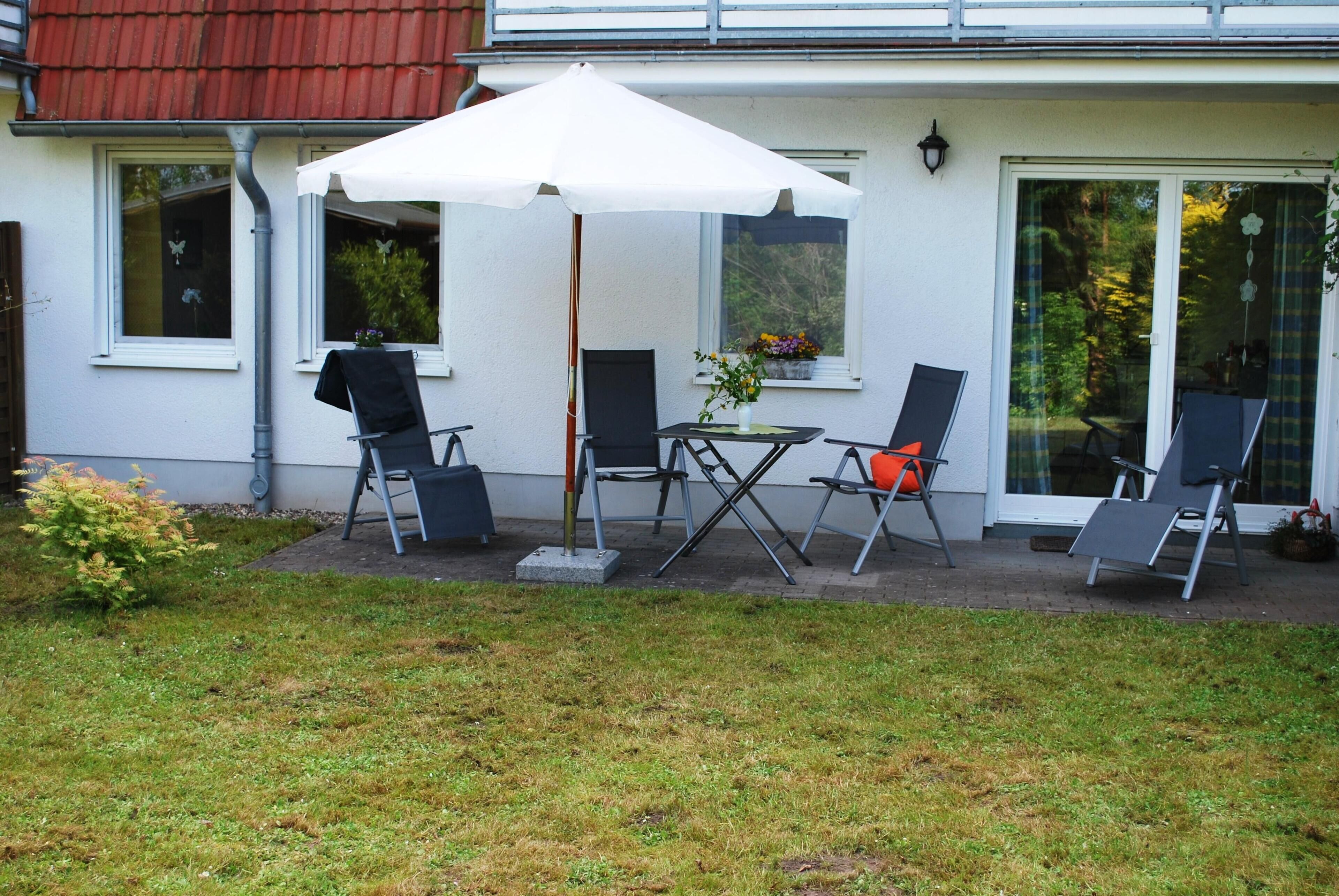 En uteplats med ett bord och stolar, ett vitt parasoll och ett hus med rött tak och stora fönster.