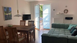 A living room with a wooden dining table, chairs, a green sofa, a television, and a sliding glass door leading to an outdoor area. - 2-zi.-fewo für 4 Personen im 1. OG Links (Graal-Müritz)