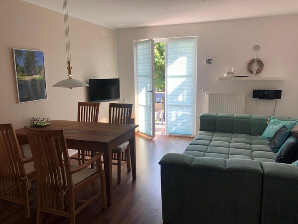 A living room with a wooden dining table, chairs, a green sofa, a television, and a sliding glass door leading to an outdoor area.