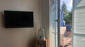 A room with a mounted television, a small wooden side table with a lantern, and a sliding glass door leading to an outdoor area with a wicker chair. - 2-zi.-fewo für 4 Personen im 1. OG Links (Graal-Müritz)
