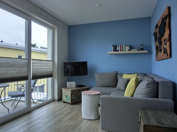 A living room with a grey sofa, a small round table, a television on a wooden stand, and a bookshelf against a blue wall.